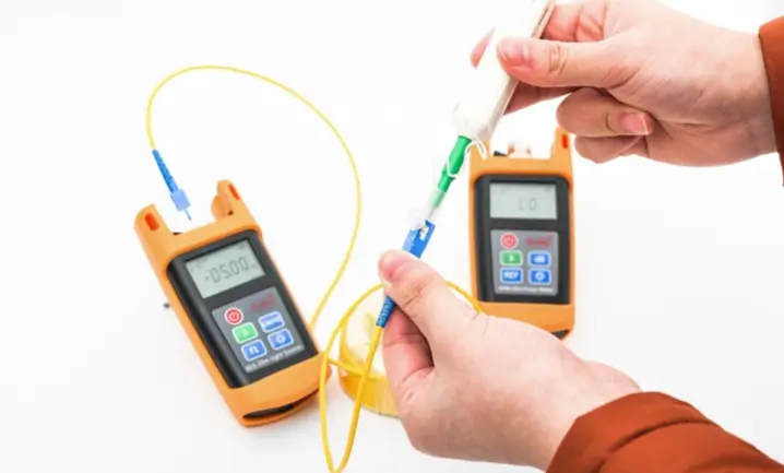 Hands connect a yellow fiber patch cord to two handheld fiber test meters during link measurement.