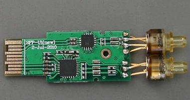 Green SFP PCB board with edge contacts, mounted chips, small components, and two optical assemblies attached at one end.
